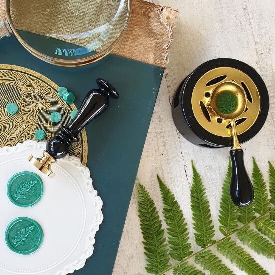 wax sealing spoon and wax melting stove in use on table with green wax sealing beads and fern wax seal stamp