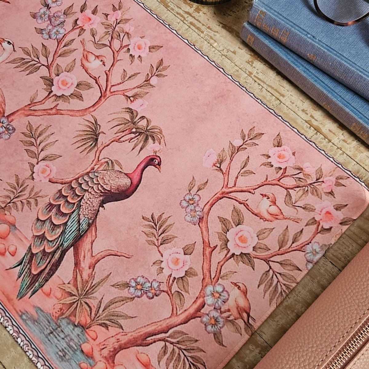 Pink desk mat with bird and floral design on a wood desk next to vintage books.