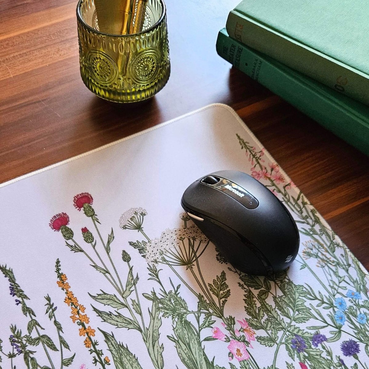 Black computer mouse on a floral-patterned mouse pad with a glass pen holder and books in the background.