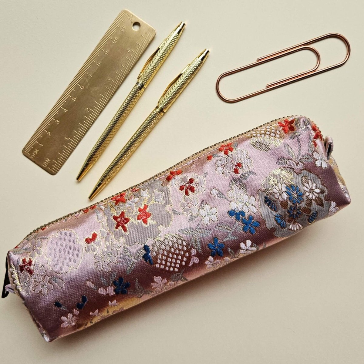 Side of pink floral embroidered pencil case with gold ruler, pens, and paperclip on a beige background