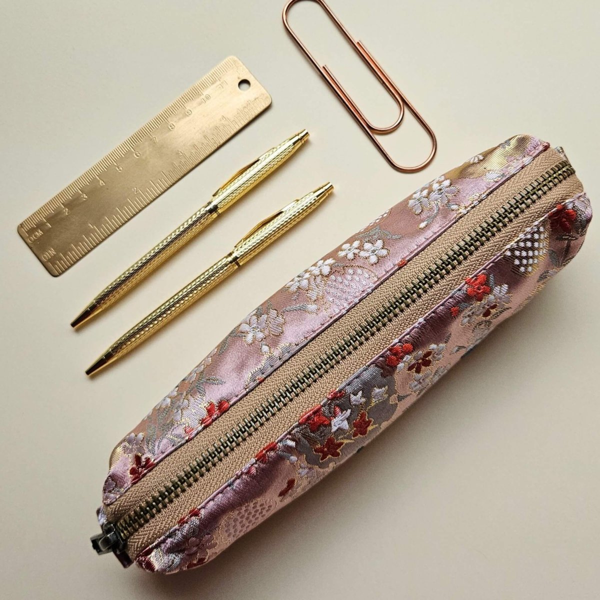 Floral pencil case with gold zipper, two gold pens, a gold ruler, and paper clips on a beige background