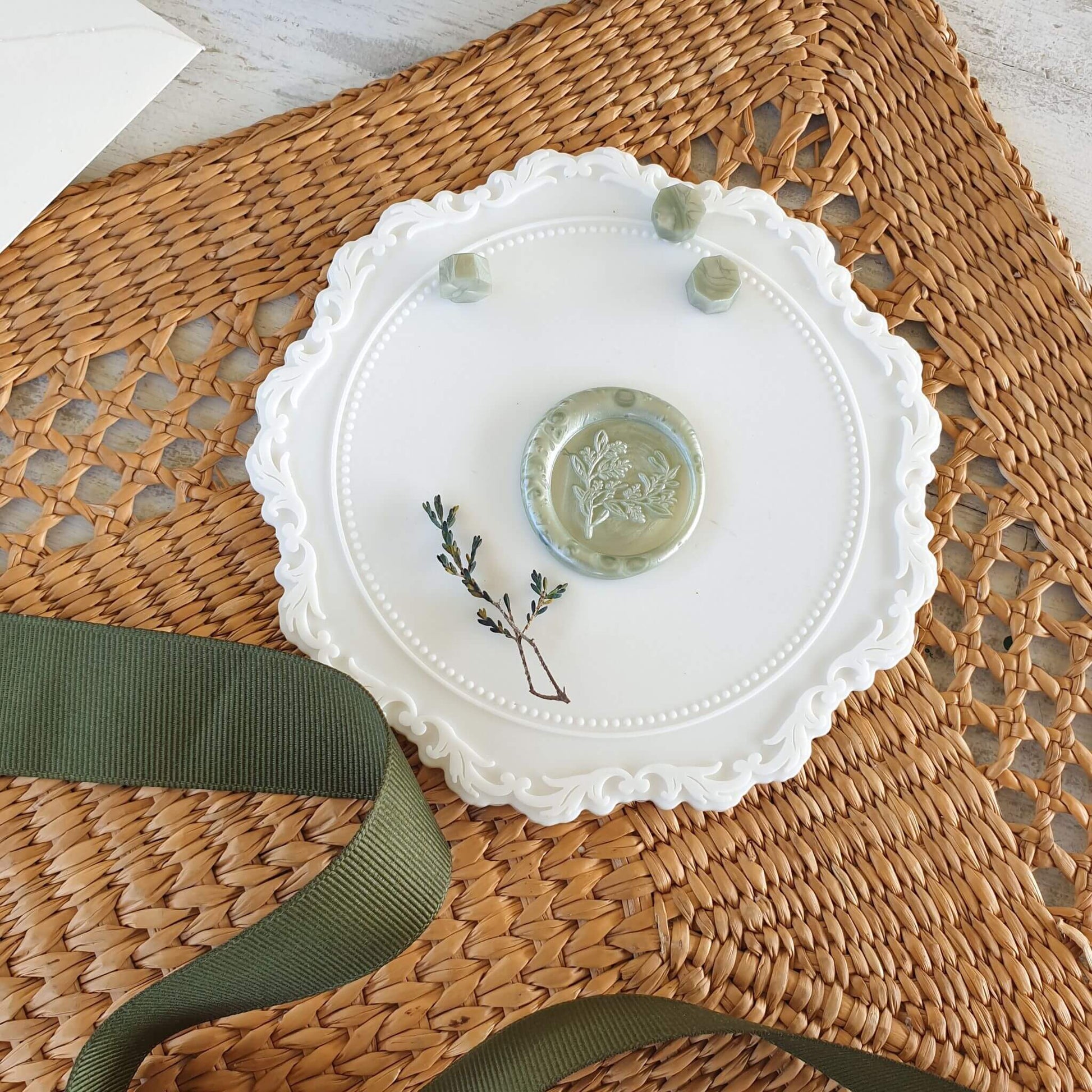 Sage green wax seal on white wax sealing mat with olive green ribbon, sage green wax sealing beads, dried flowers and rattan mat