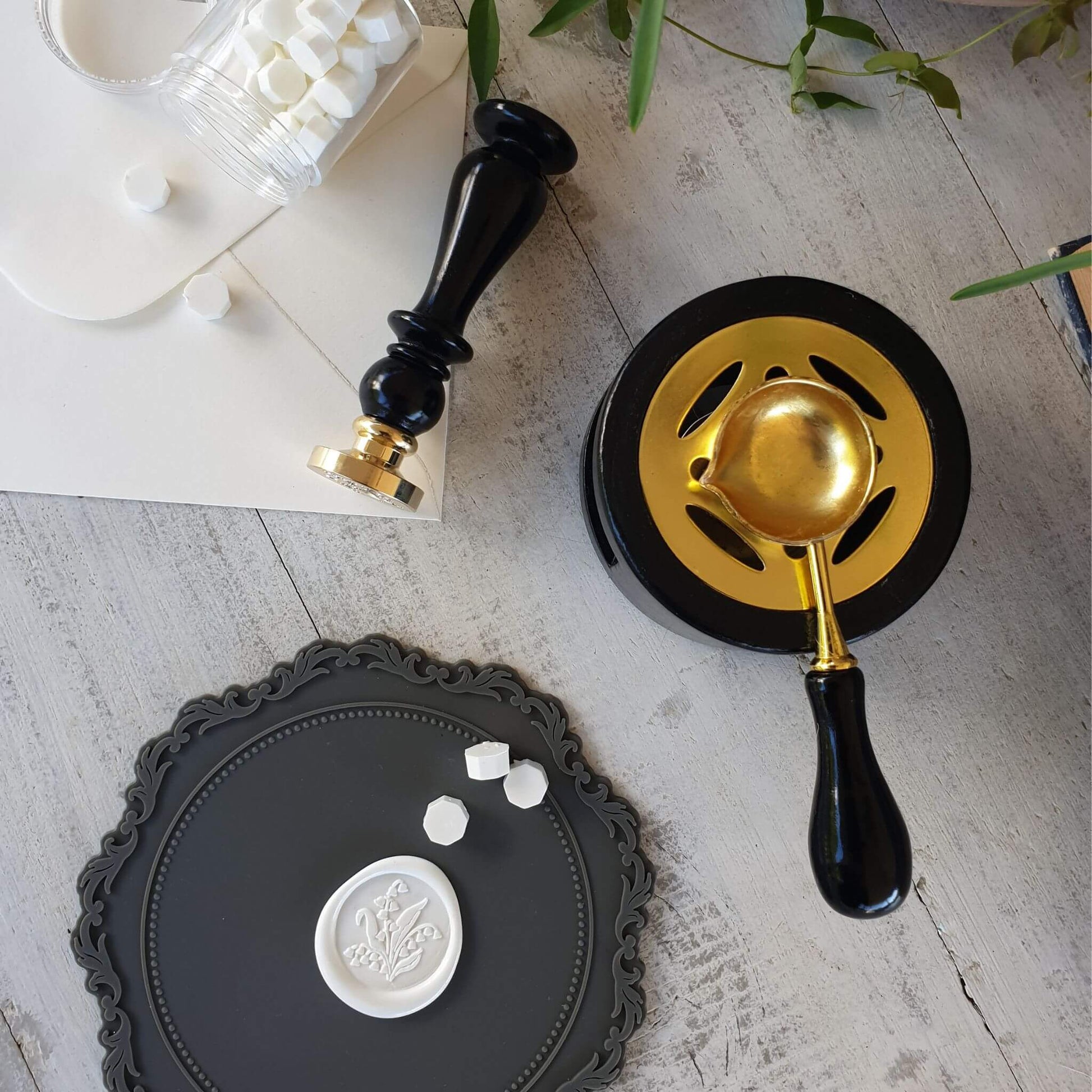 flatlay of black and gold wax melter stove in use on table with wax seal stamp, white floral wax seal, white wax sealing beads and grey silicone wax sealing mat