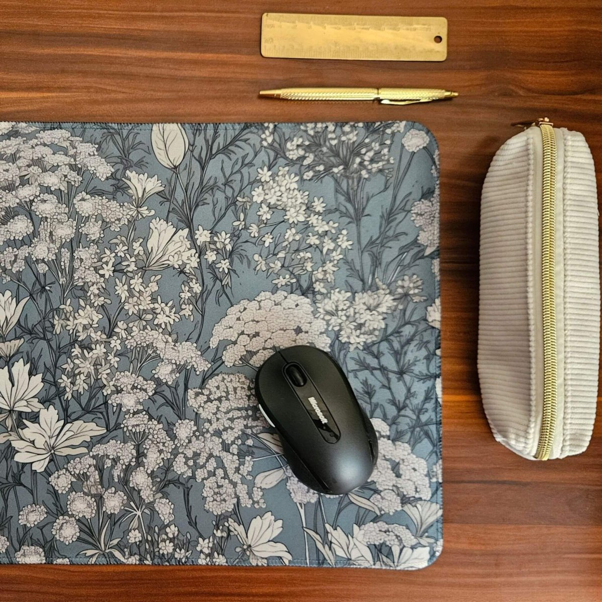 Floral mouse pad with a computer mouse, gold pen, and white pouch on a wooden desk.