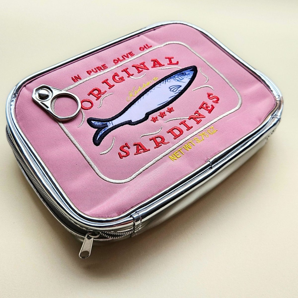 Pink sardine tin-shaped pouch with fish illustration on a beige background