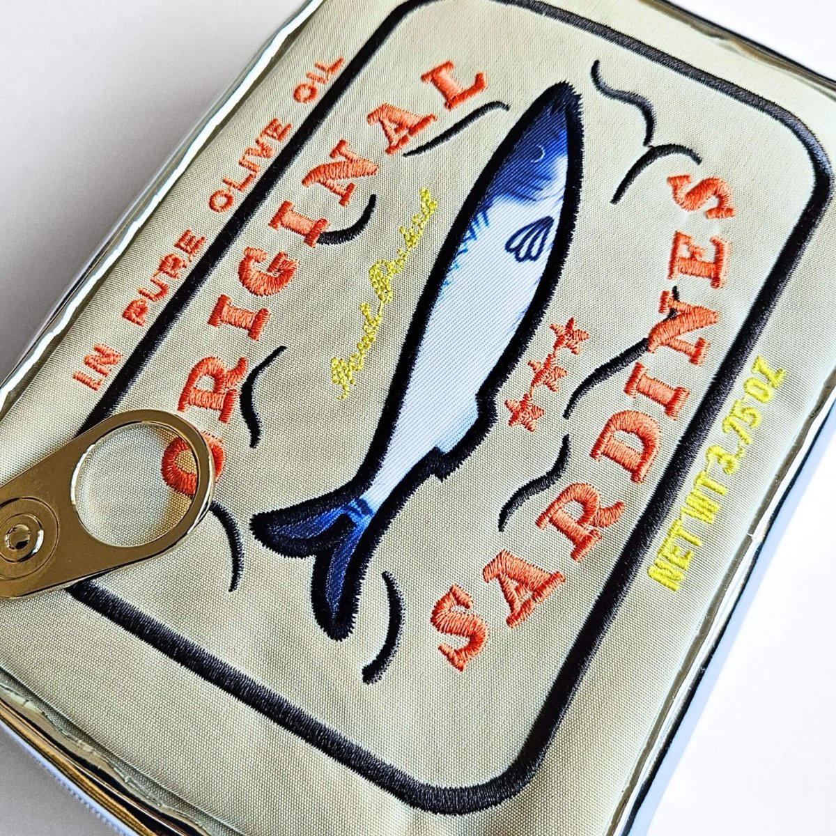 Sardine can-shaped pouch with sardine illustration and text on a beige background