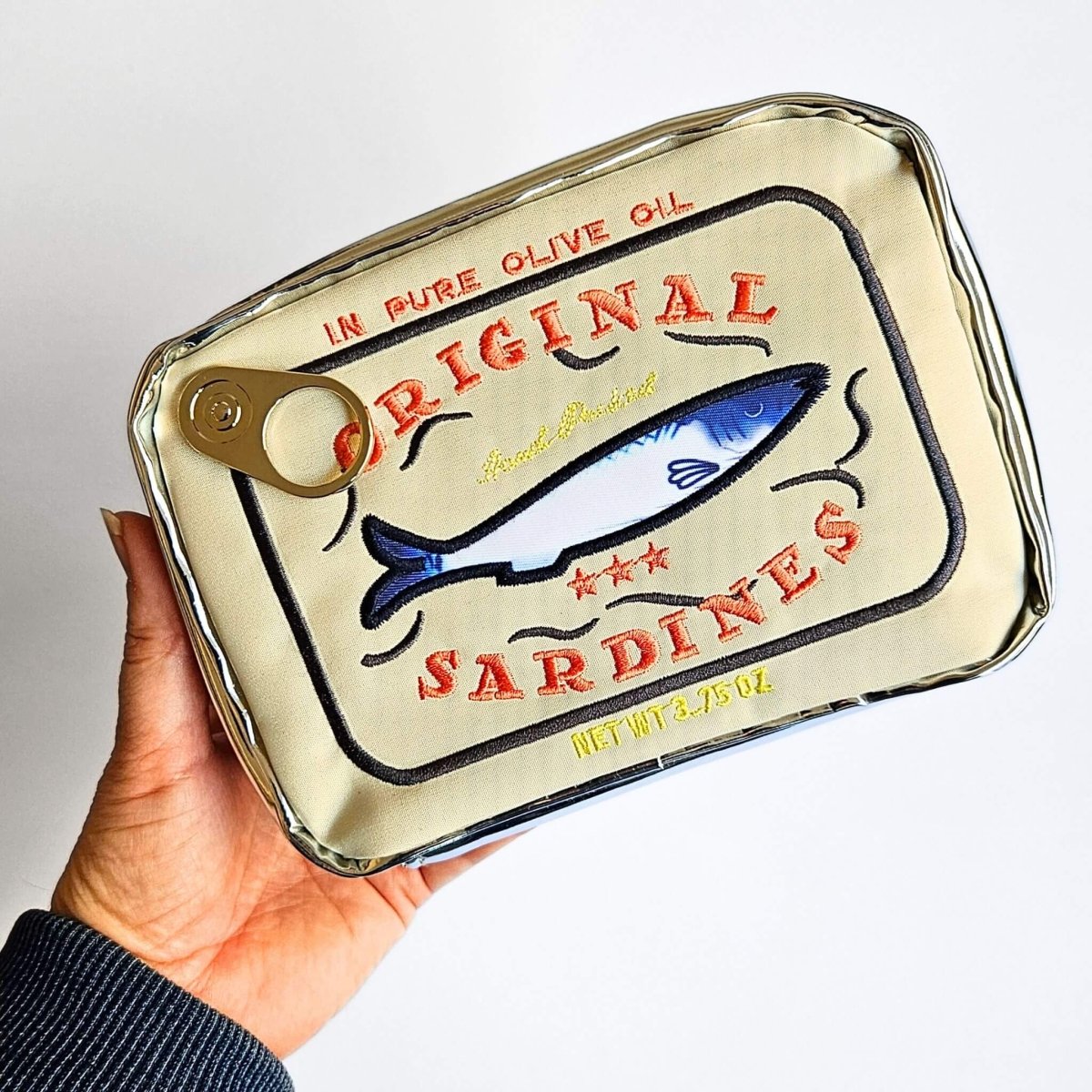 Hand holding a sardine can with 'Original Sardines' label on a light gray background