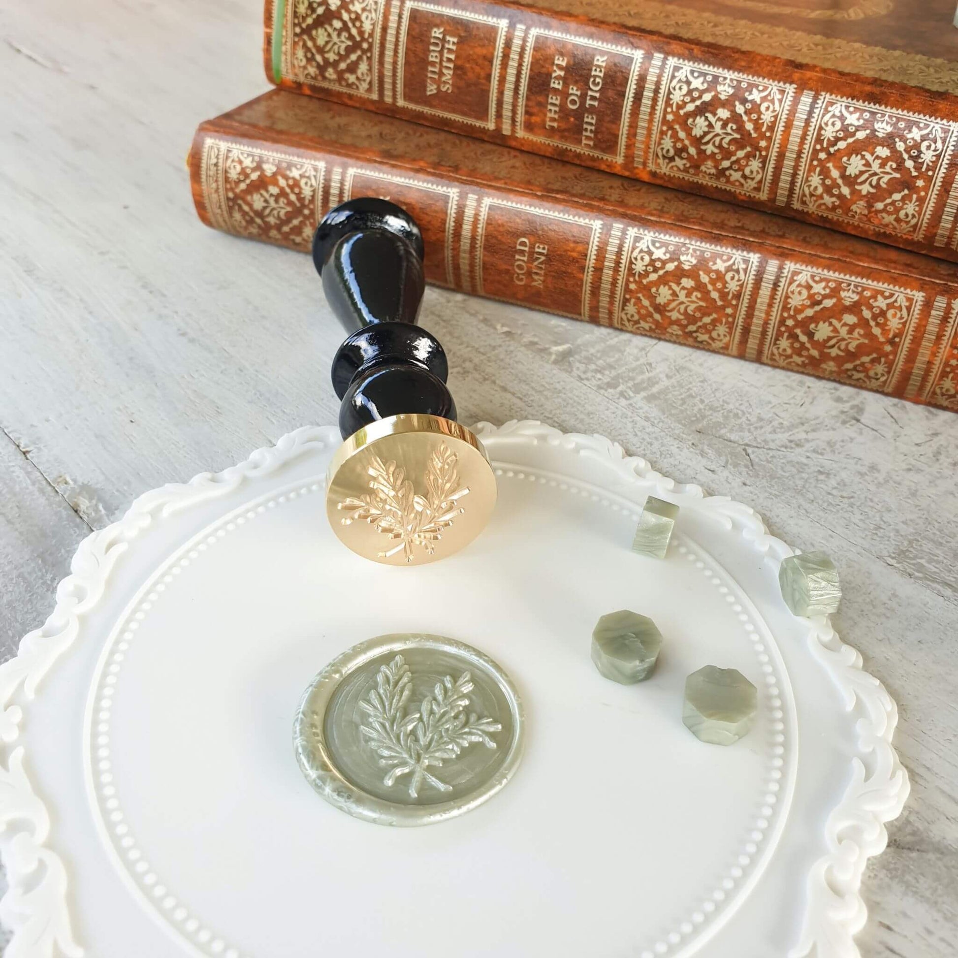 white wax sealing mat in use on table with sage green foliage wax seal, wax seal stamp and sage green wax sealing beads next to vintage books